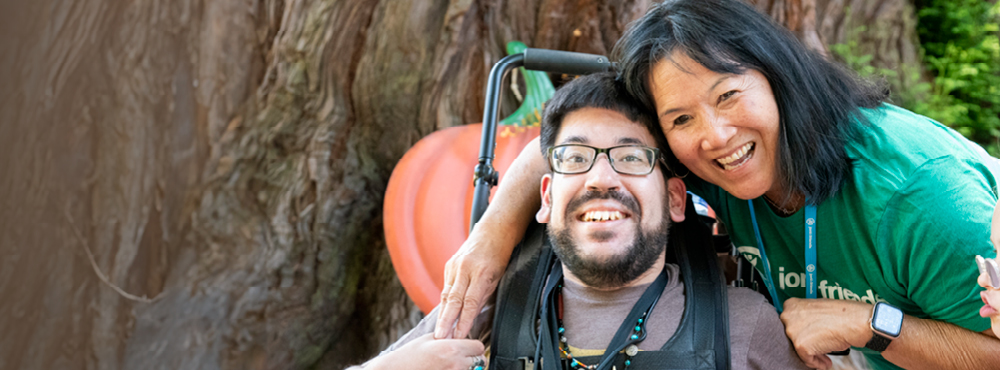 A man in a wheelchair and a woman are hugging each other and smiling at the camera, with a large tree bark in the background.