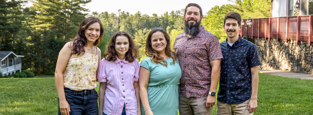 A family of five smiling and posing for the camera against a backdrop of lush green grass and trees.