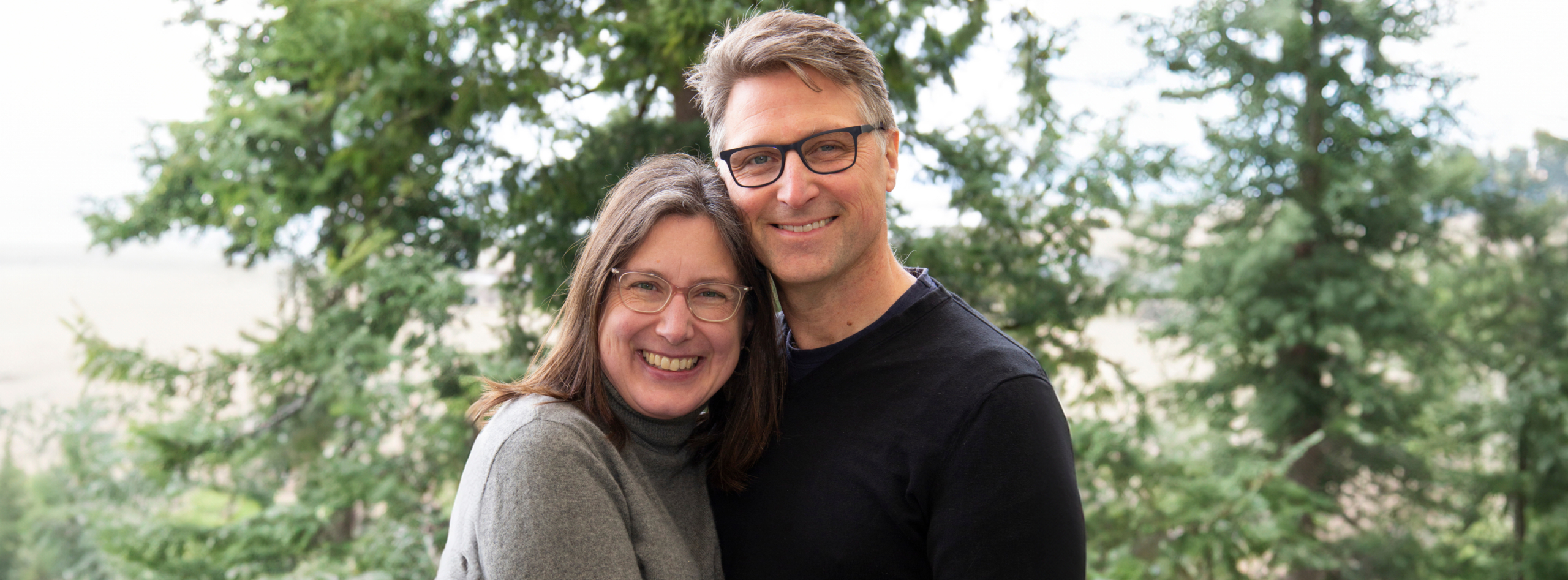 A couple smiling at the camera against a backdrop of blurred trees.