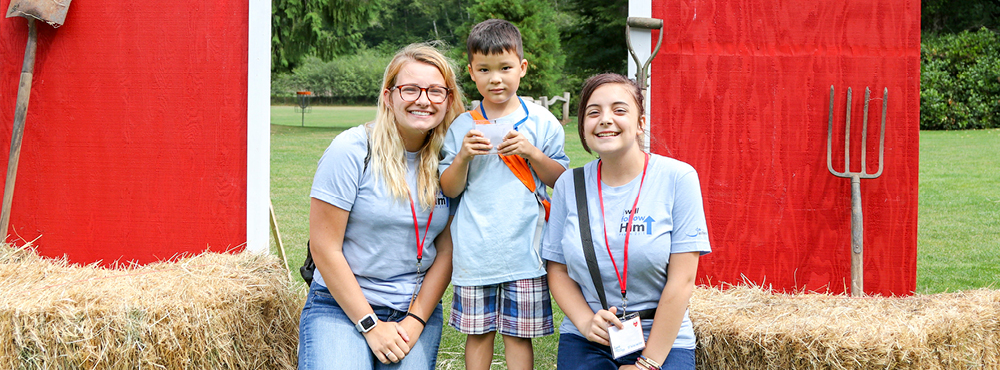 On the left, an enthusiastic woman volunteer and on the right, another woman smiling beside a cheerful boy in the middle, all posing for the camera against a backdrop of a charming hay barn, capturing a moment of joy.