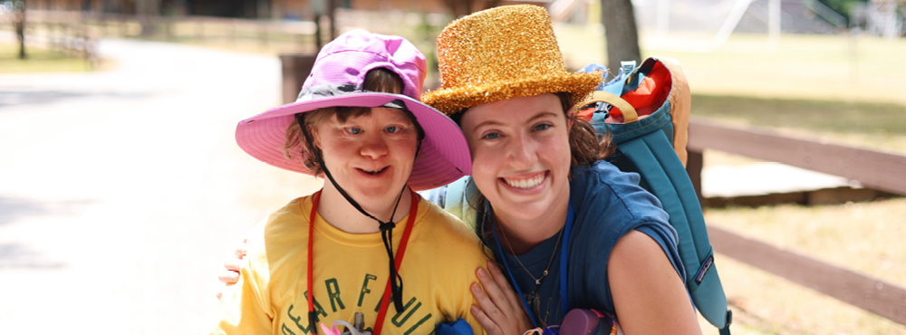 A child wearing a yellow shirt and a pink hat, along with a volunteer dressed in a blue shirt and a golden hat, both smiling and posing cheerfully for the camera.