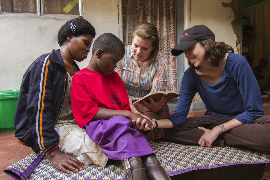 A little boy sits on his mother's lap, holding hands with a volunteer, while another volunteer reads from an open Bible.