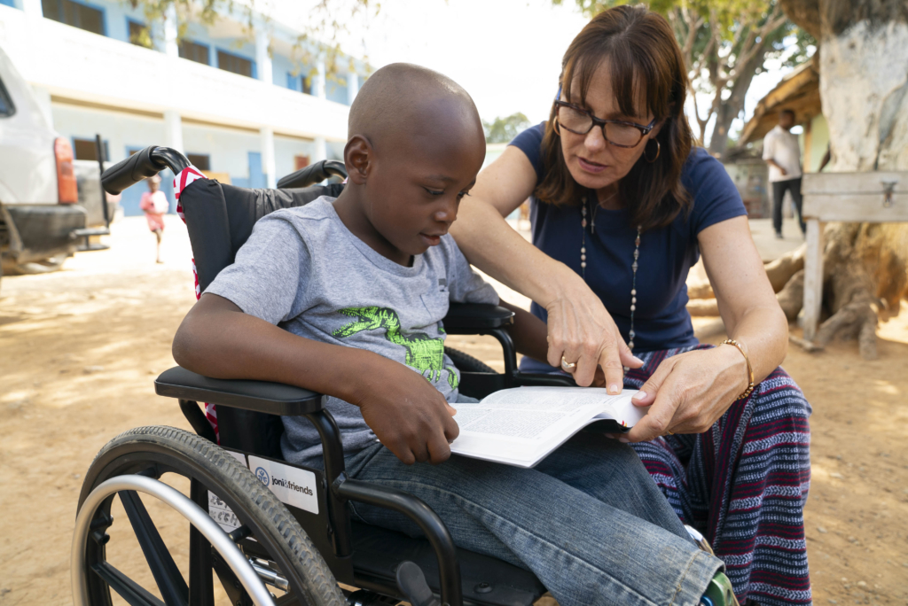 A boy in a wheelchair reads the Bible in his lap while a volunteer sits beside him, pointing to a scripture.