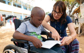 A boy in a wheelchair reads the Bible in his lap while a volunteer sits beside him, pointing to a scripture.