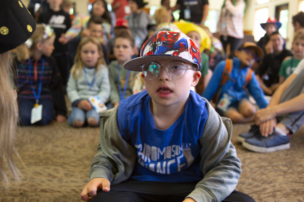 Colton sits on a carpet, wearing a Spiderman cap, a blue shirt, and a grey jacket, looking focused. In the background, other children are seated on the carpet, slightly blurred.