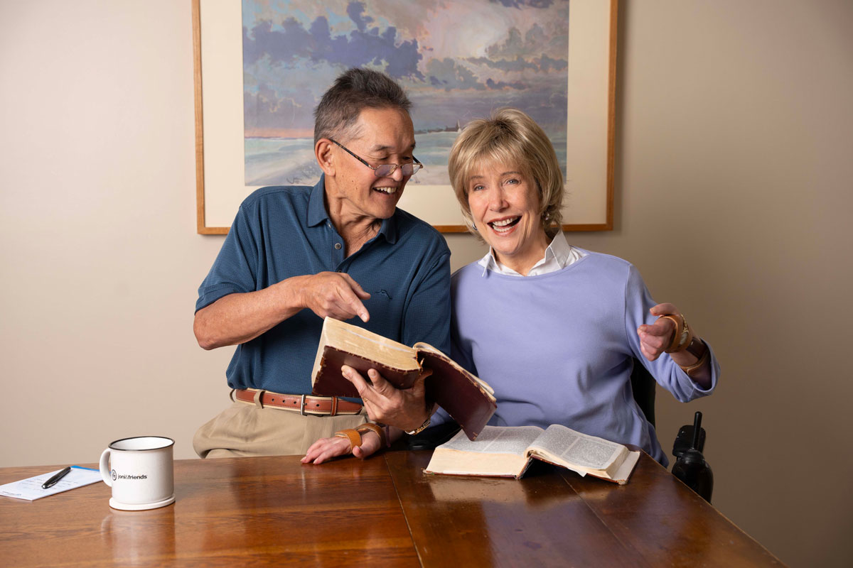 Ken holds a Bible and points at it while looking at Joni, who is smiling and looking at the camera with an open Bible on the table.