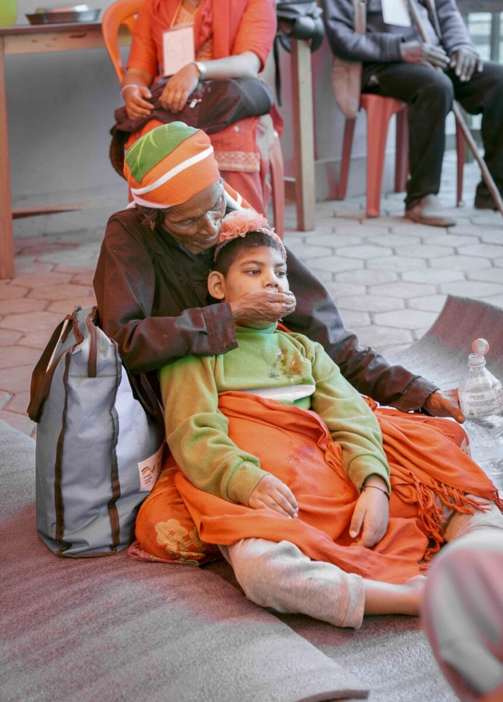 A child leans against a woman seated on the floor, who is gently feeding her.