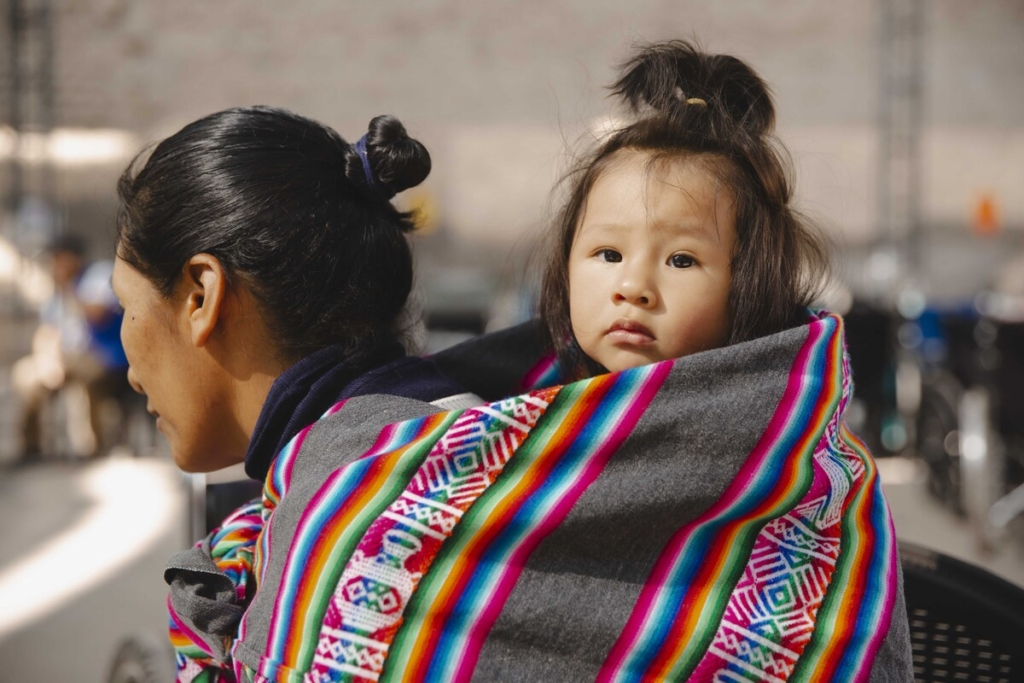 A child wrapped in a colorful cloth behind her mother.