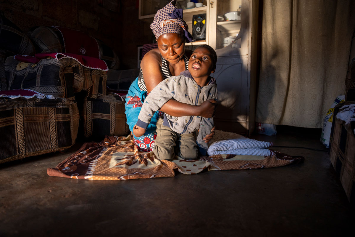 Chisomo, a child with a disability, is sitting on the floor while his mother struggles to lift him up. The scene highlights the mother's effort and determination.