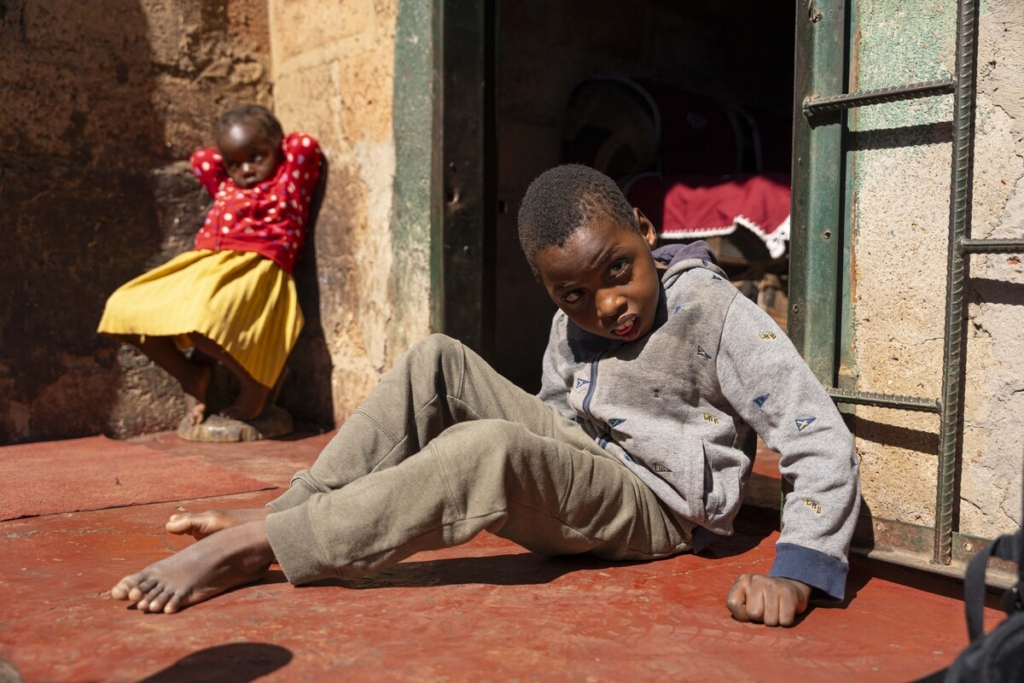 Chisomo sitting on a red floor in dirty clothes, with his sister in the background.