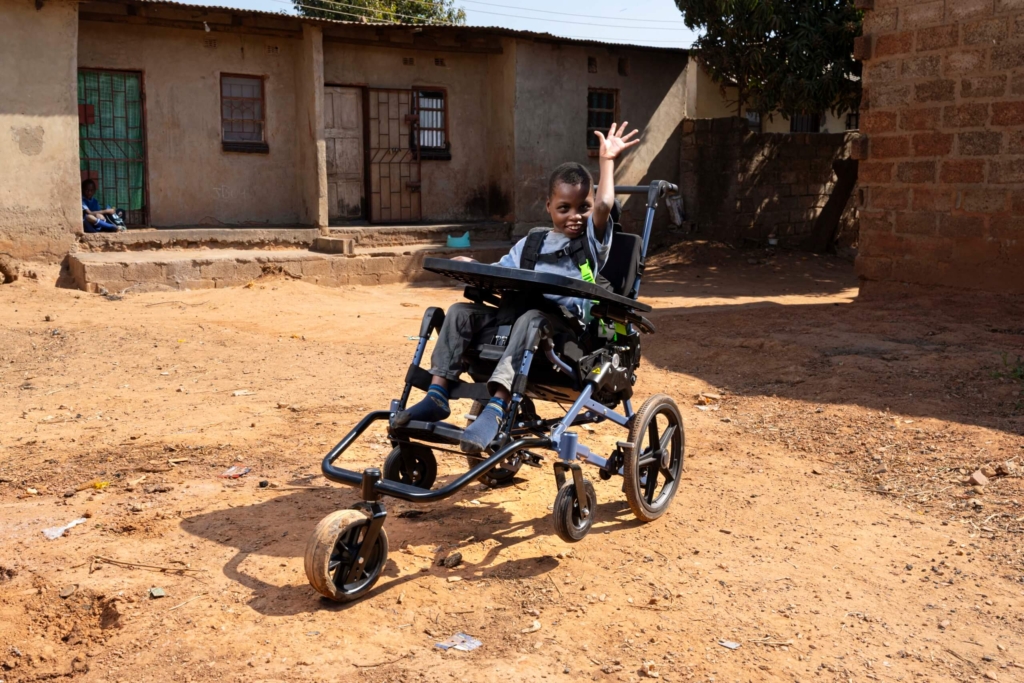 Chisomo smiling and waving at the camera from his new Cub wheelchair, radiating joy and excitement.