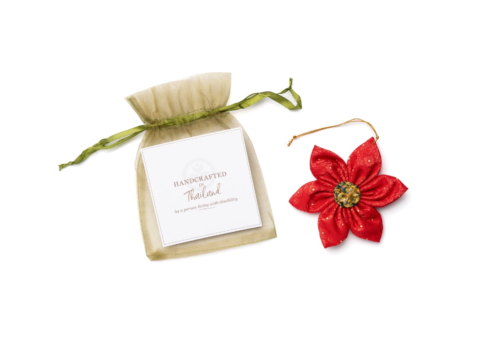 The Poinsettia ornament is next to its gift pouch, which has a card that states, "Handcrafted in Thailand by a person living with disability."