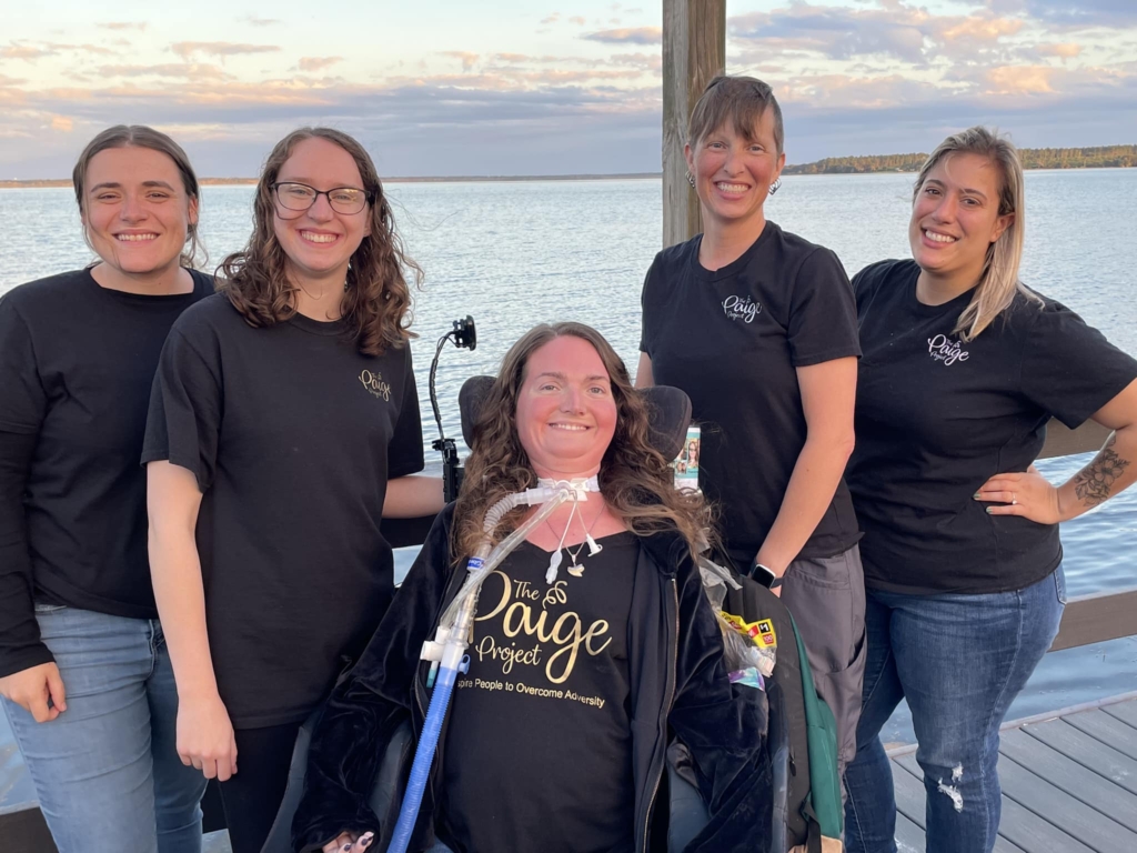 Paige Snedeker, in her wheelchair and wearing a black t-shirt with golden text that reads "The Paige Project Inspiring People to Overcome Adversity," in between among four women in black shirts featuring a small The Paige Project logo.