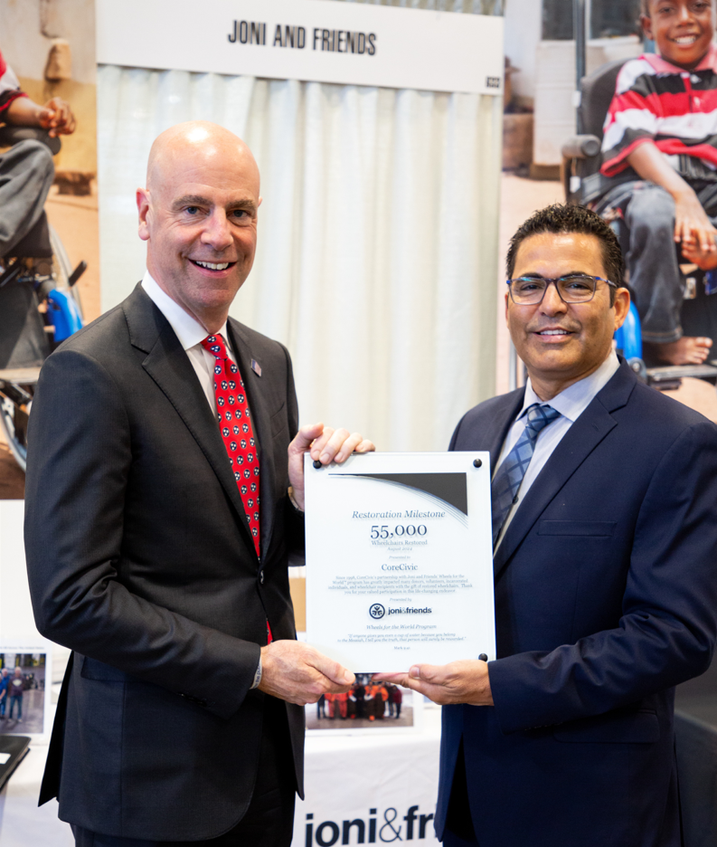 Damon T. Hininger, President and CEO of CoreCivic, and Paul Dorthalina, Director of Wheels for the World at Joni and Friends, are smiling and posing for the camera while holding an award together.