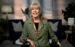 Joni Eareckson Tada, wearing a green denim jacket and a scarf, is smiling at the camera. The background features a living room setting.