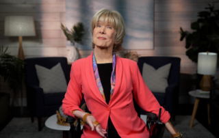 Joni Eareckson Tada, wearing a vibrant light red blazer over a black top and a colorful scarf, is smiling as she raises her left arm for the camera. The background features a softly blurred living room setup, adding a cozy atmosphere to the image.