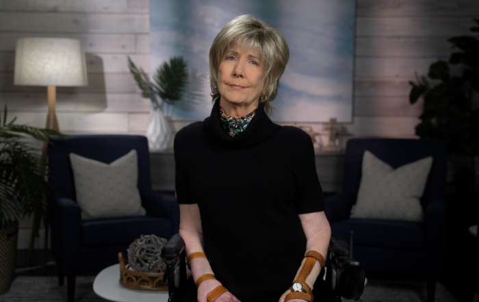 Joni Eareckson Tada smiles for the camera, wearing a black blouse and colorful scarf, with a blurred living room backdrop.