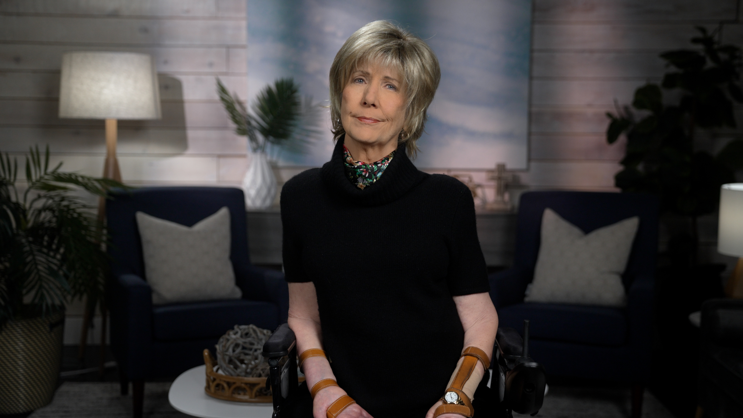 Joni Eareckson Tada smiles for the camera, wearing a black blouse and colorful scarf, with a blurred living room backdrop.