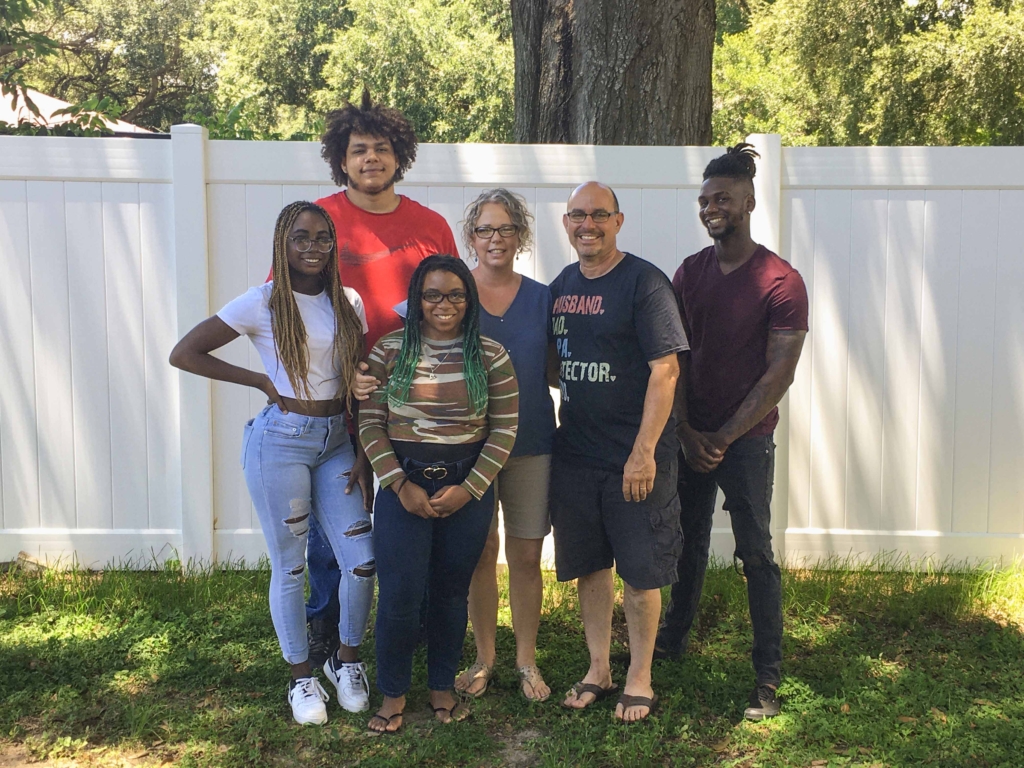 A beautiful family picture of Sara Pueblo's family, including Esther, her three siblings, and her husband David, standing on green grass in front of a white fence.