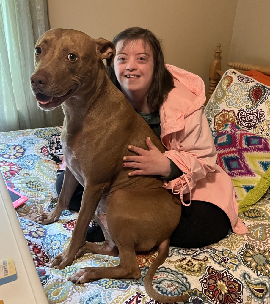 Joana sits on her bed, smiling joyfully while holding her big brown dog, who is also displaying a happy expression. The cozy bedroom setting adds to the cheerful atmosphere.