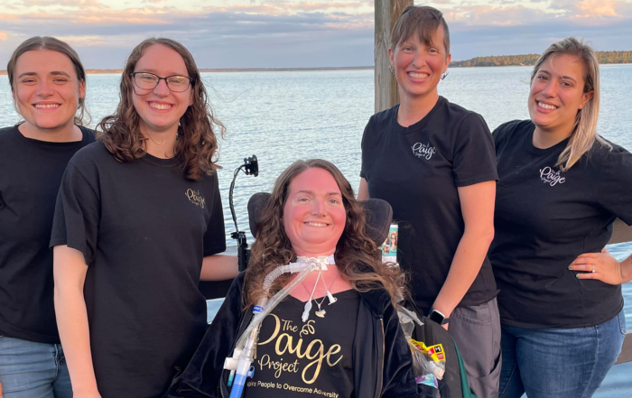 Paige Snedeker, in her wheelchair and wearing a black t-shirt with golden text that reads "The Paige Project Inspiring People to Overcome Adversity," in between among four women in black shirts featuring a small The Paige Project logo.