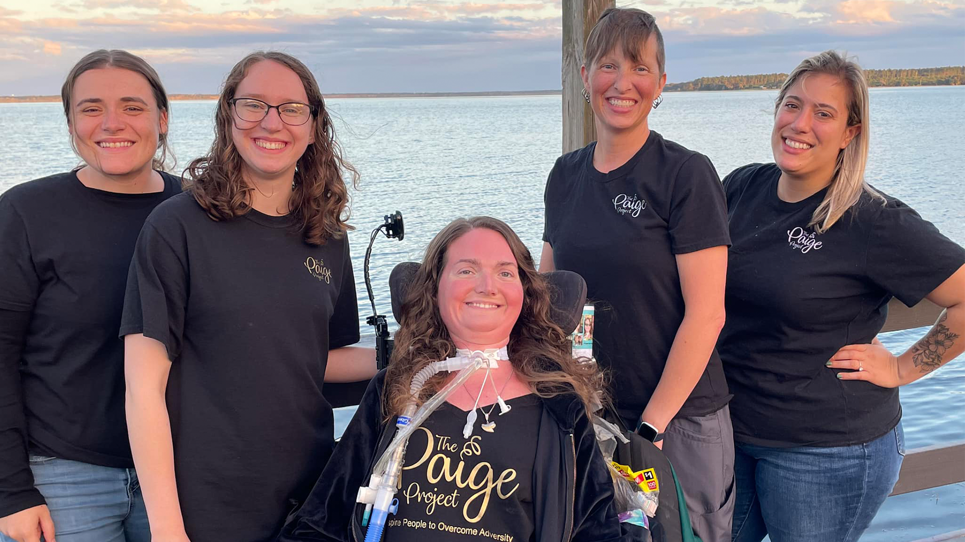 Paige Snedeker, in her wheelchair and wearing a black t-shirt with golden text that reads "The Paige Project Inspiring People to Overcome Adversity," in between among four women in black shirts featuring a small The Paige Project logo.