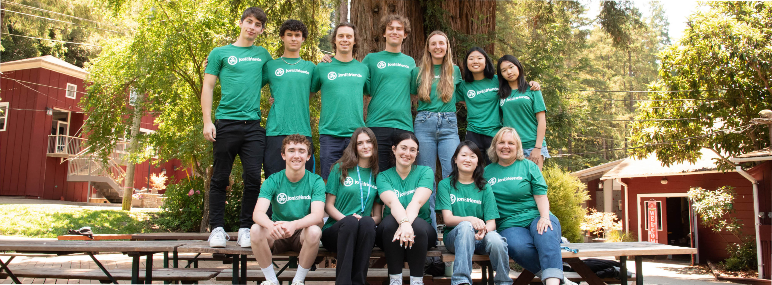 A group of interns at Joni and Friends, wearing green t-shirts with the Joni and Friends logo. Five interns are seated at a wooden table, while seven are standing behind them. In the background, a large tree towers over the scene, surrounded by quaint red houses and lush greenery.