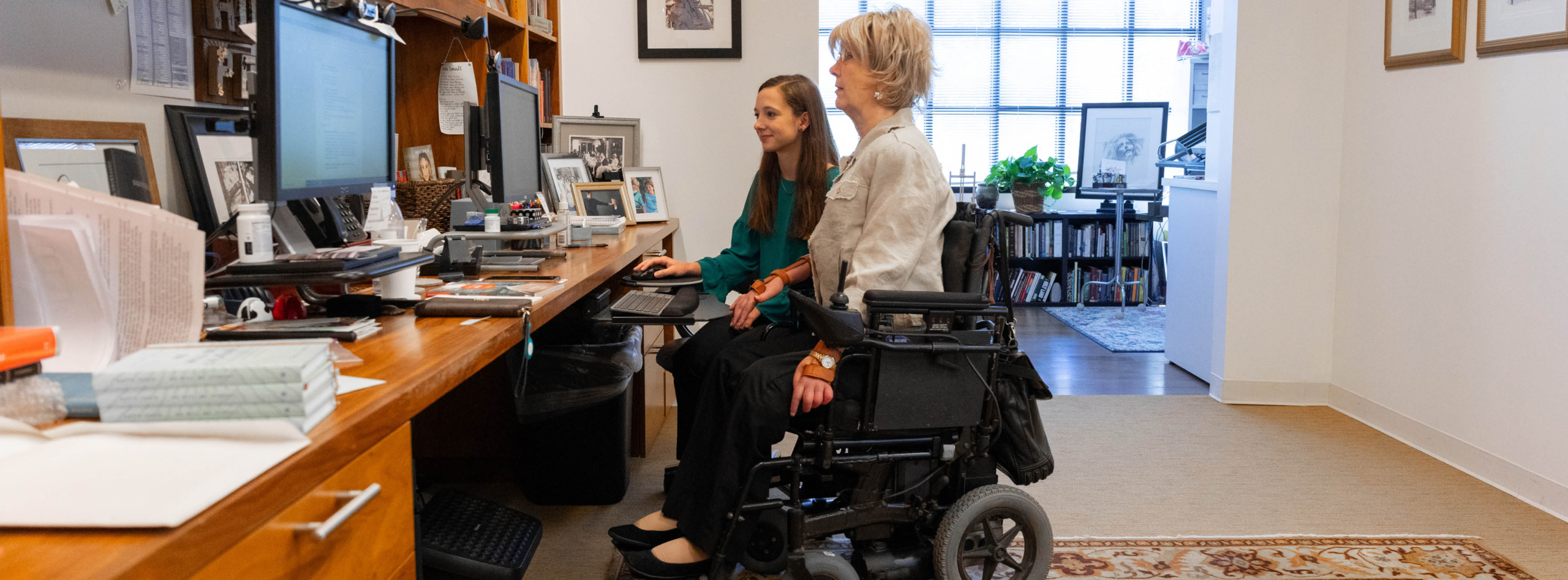 Side view of Joni Eareckson Tada in her wheelchair, looking at a computer monitor, next to an intern who is holding the mouse and also looking at the screen.