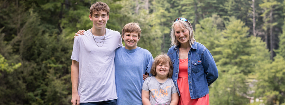 A joyful family portrait featuring two boys on the left, a girl, and their mom, all looking so joyful with huge smiles on their faces, set against a backdrop of lush green tall trees.