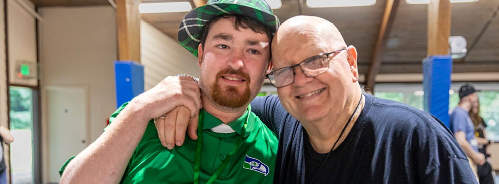 Two men are smiling and posing for the camera. One man wears a plaid hat and a green shirt, while the other wears a dark blue shirt and glasses. They both look very happy.