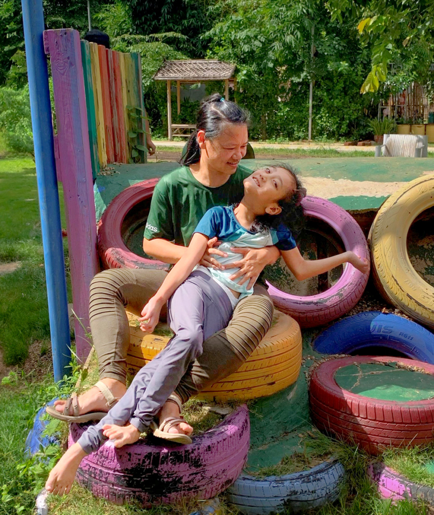 A heartwarming photo of Yupin carrying Air in her lap as they sit on colorful tires against a lush green background. 