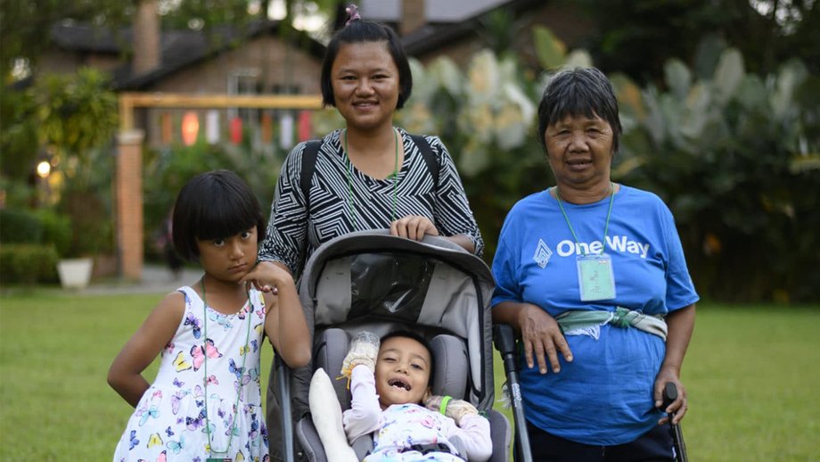 Yupin behind Air in her stroller, with Praewa on the left and another woman with a cane, all smiling and posing for the camera in a grassy area with a slightly blurry background.