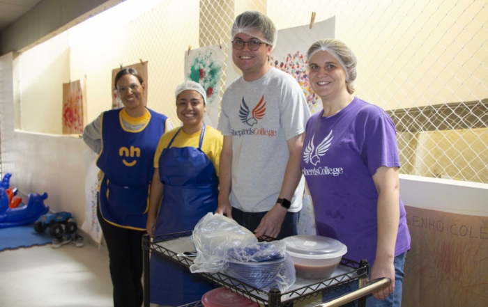 Matthew Gilliland is wearing a hairnet and a t-shirt with "Shepherd's College" on it, posing with three other interns. In front of them is a cart with plates and food.