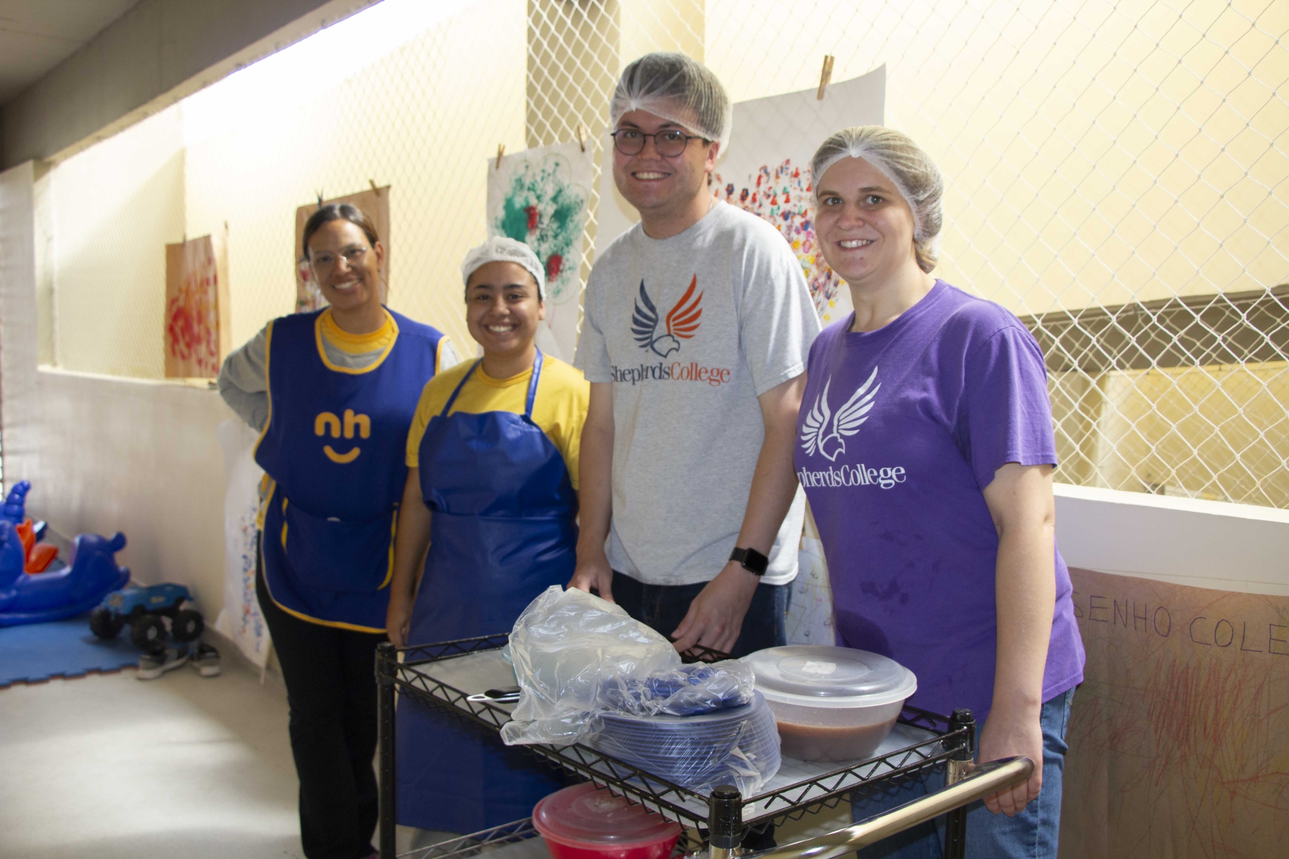 Matthew Gilliland is wearing a hairnet and a t-shirt with "Shepherd's College" on it, posing with three other interns. In front of them is a cart with plates and food.