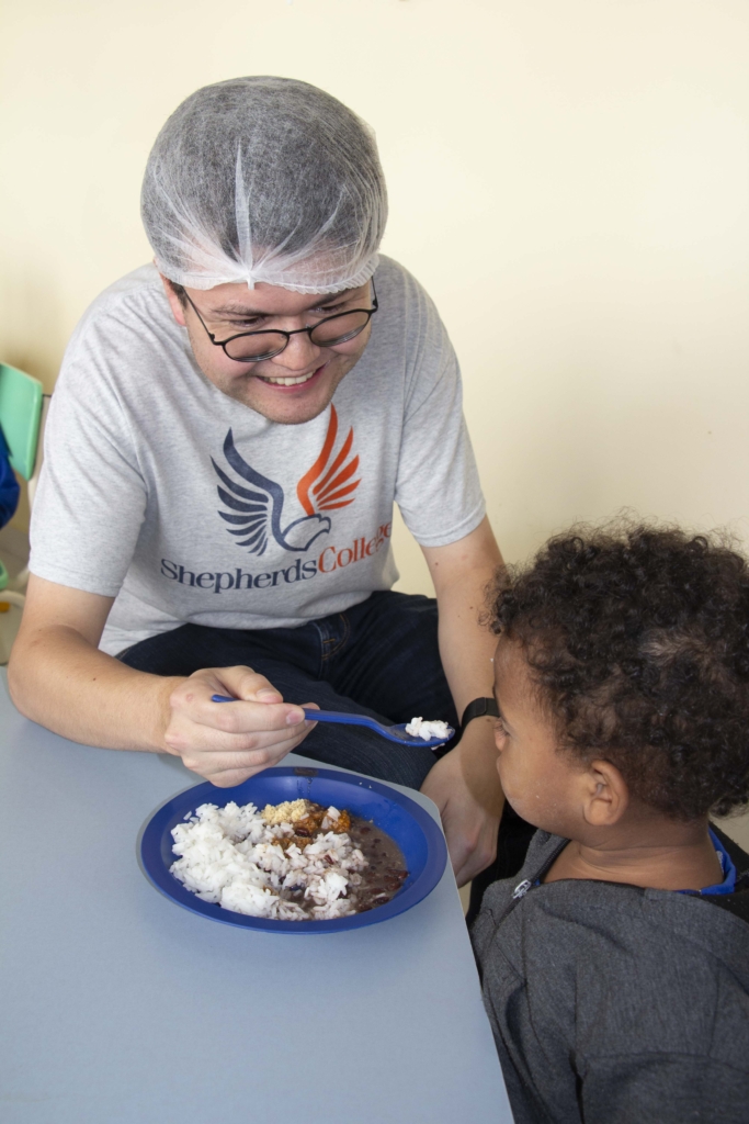 Matthew Gilliland is wearing a hairnet and a t-shirt with "Shepherd's College" on it, holding a spoon with rice and feeding a young child, with a plate on the table in front of them.