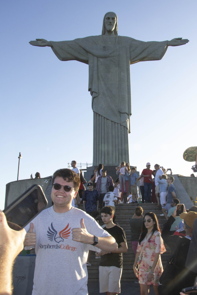 Matthew Gilliland is posing for the camera with both hands giving a thumbs up, wearing a Shepherd's College t-shirt, in front of the Christ the Redeemer statue in Brazil, with a crowd of people behind him.