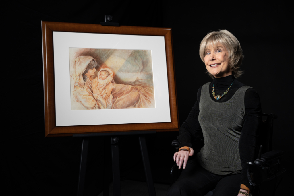 Joni is smiling and sitting in her wheelchair next to her artwork titled "The Nativity."