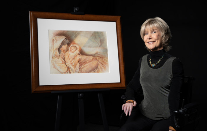 Joni is smiling and sitting in her wheelchair next to her painting titled "The Nativity."