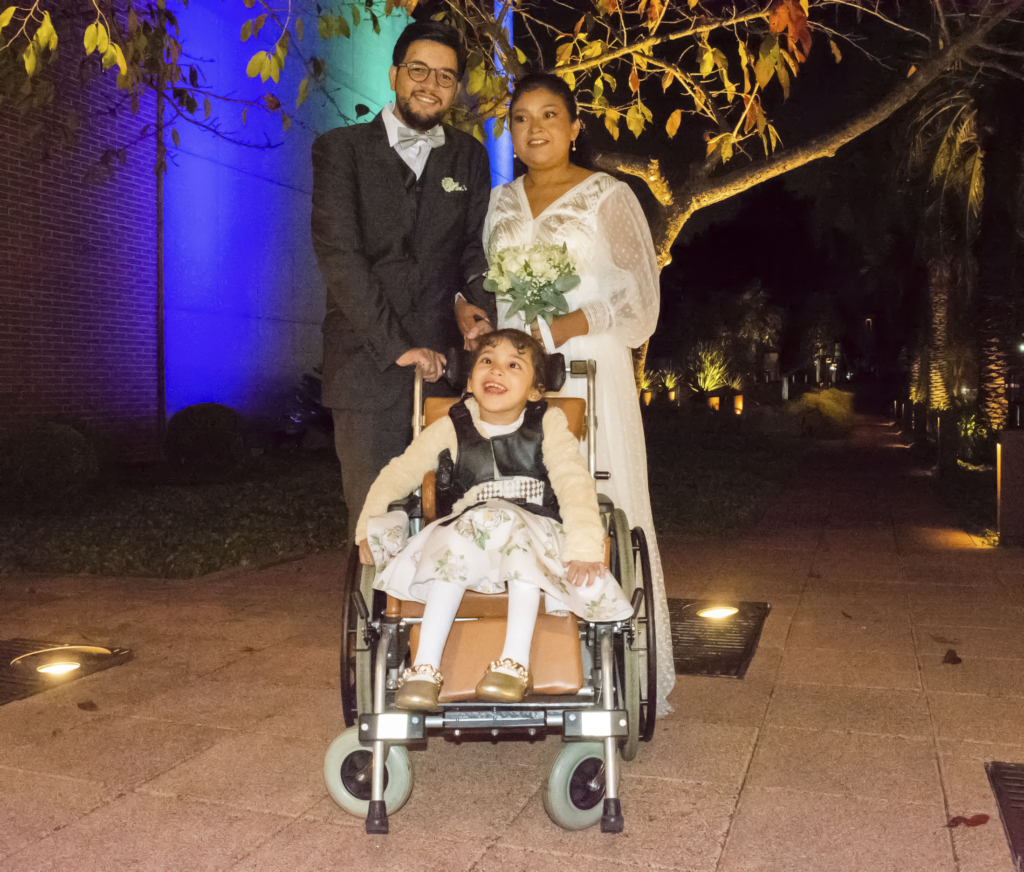 A picture of Andrew and Alba standing together behind Sofia's wheelchair on their wedding day. Andrew is holding the push handle of Sophia's wheelchair, while Alba, wearing her white wedding gown and holding a bouquet of flowers, is holding Andrew's hand with her other hand. Sofia looks happy and excited, wearing her dress and sitting in her wheelchair.