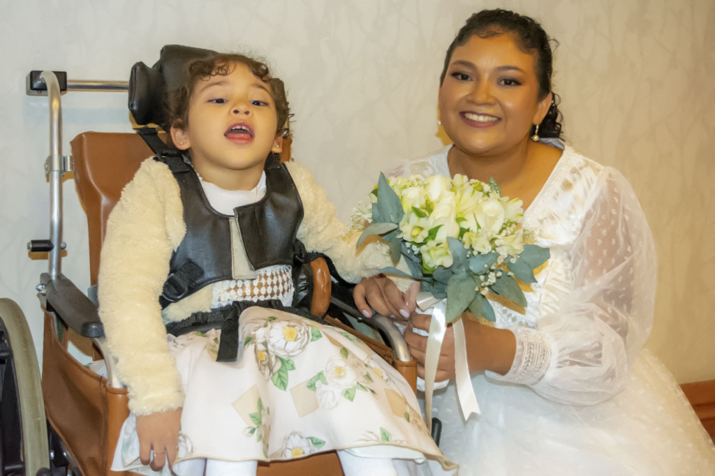 Sofiia in her wheelchair, wearing a beautiful flowery dress, next to Alba, who is wearing her white wedding gown and holding a bouquet of flowers. Both are posing and smiling for the camera.