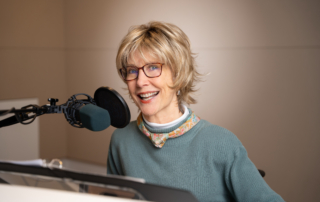 Joni smiles at the camera in a radio studio, posing with a microphone in front of her. She wears a sweater, a colorful floral scarf, and eyeglasses.