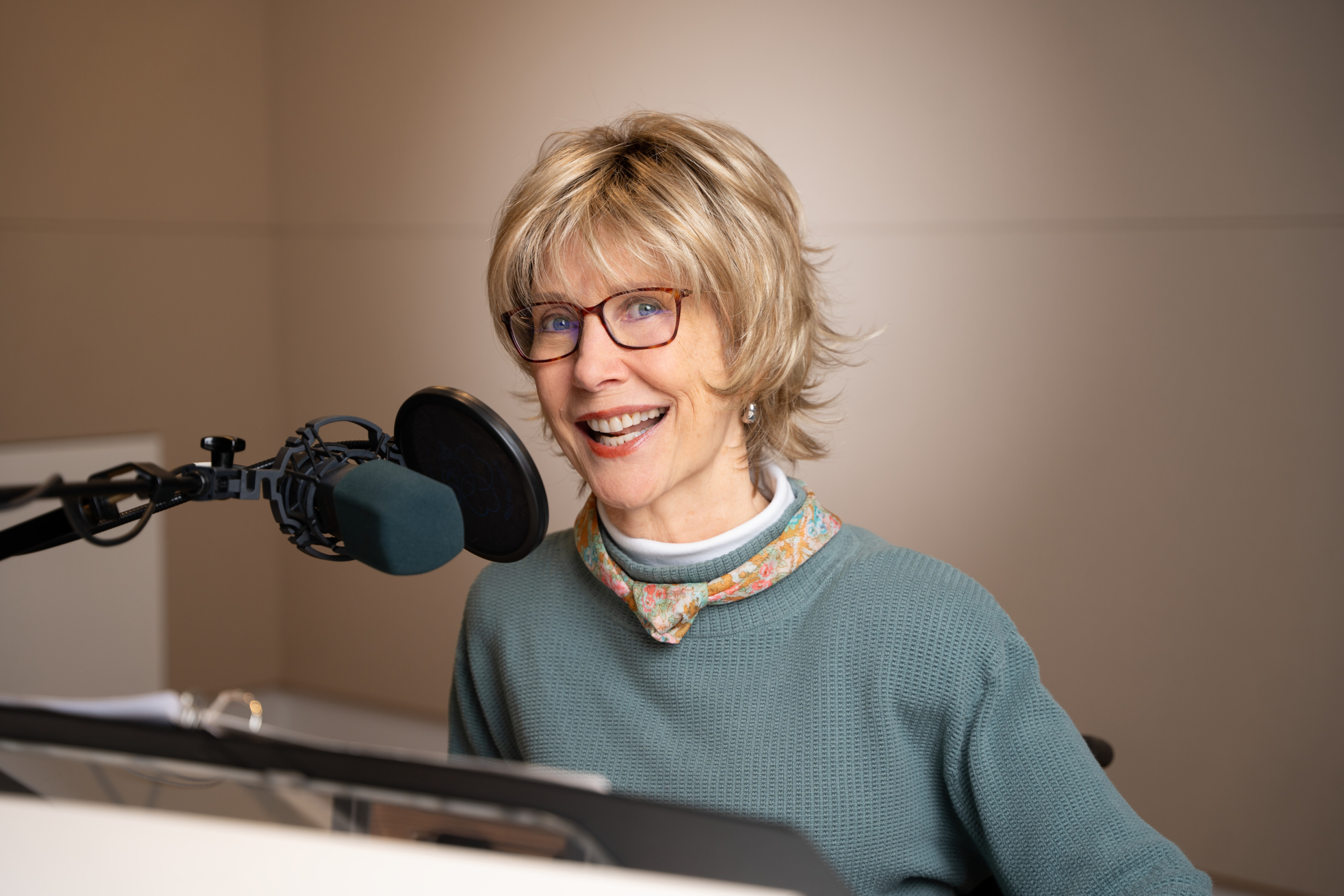 Joni smiles at the camera in a radio studio, posing with a microphone in front of her. She wears a sweater, a colorful floral scarf, and eyeglasses.