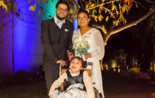 A picture of Andrew and Alba standing together behind Sophia's wheelchair on their wedding day. Andrew is holding the push handle of Sophia's wheelchair, while Alba, wearing her white wedding gown and holding a bouquet of flowers, is holding Andrew's hand with her other hand. Sophia looks happy and excited, wearing her dress and sitting in her wheelchair.