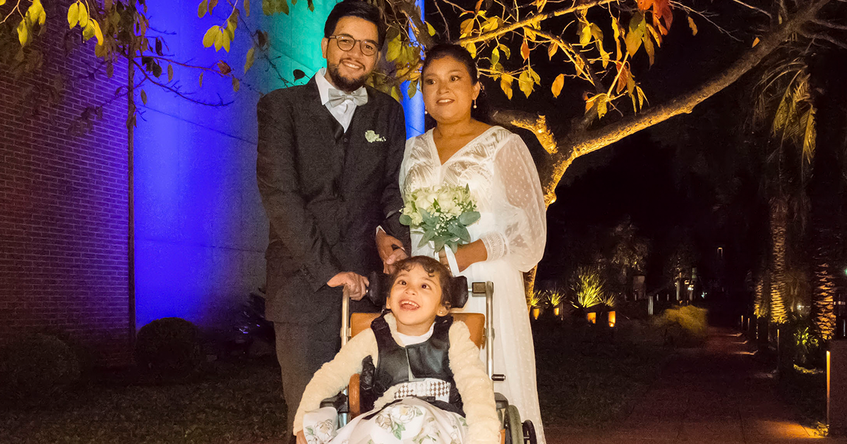 A picture of Andrew and Alba standing together behind Sophia's wheelchair on their wedding day. Andrew is holding the push handle of Sophia's wheelchair, while Alba, wearing her white wedding gown and holding a bouquet of flowers, is holding Andrew's hand with her other hand. Sophia looks happy and excited, wearing her dress and sitting in her wheelchair.