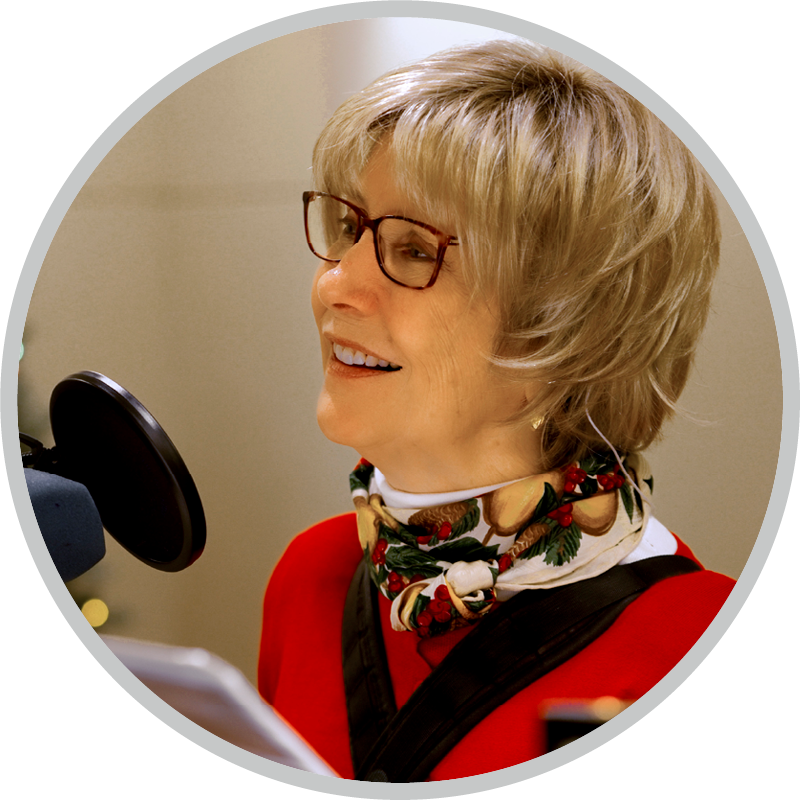 Side view shot of Joni wearing a red long-sleeve shirt, a colorful scarf, and eyeglasses, with a microphone in front of her.