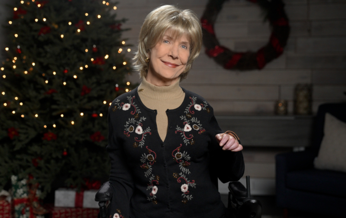 Joni smiles for the camera, wearing a light brown turtleneck under a black long-sleeve sweater with embroidered flowers and leaves. The background features a cozy living room with a Christmas tree and wrapped gifts.