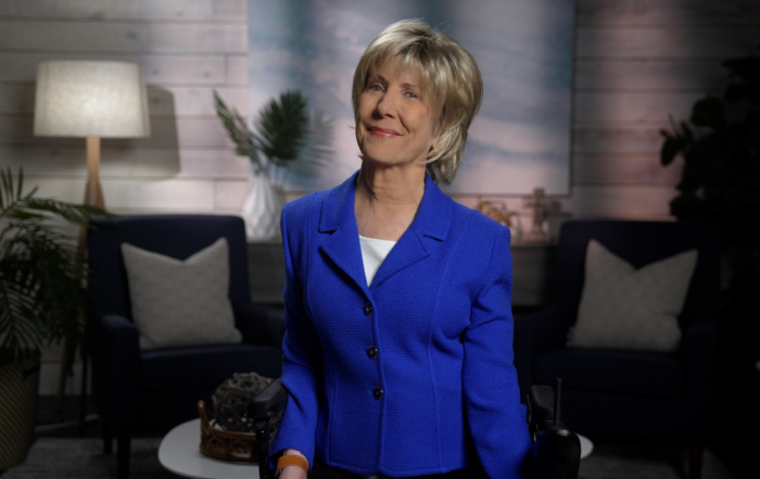 Joni Eareckson Tada is smiling for the camera, wearing a royal blue jacket over a white top. She is positioned in front of a softly blurred background that features a cozy living room setup.