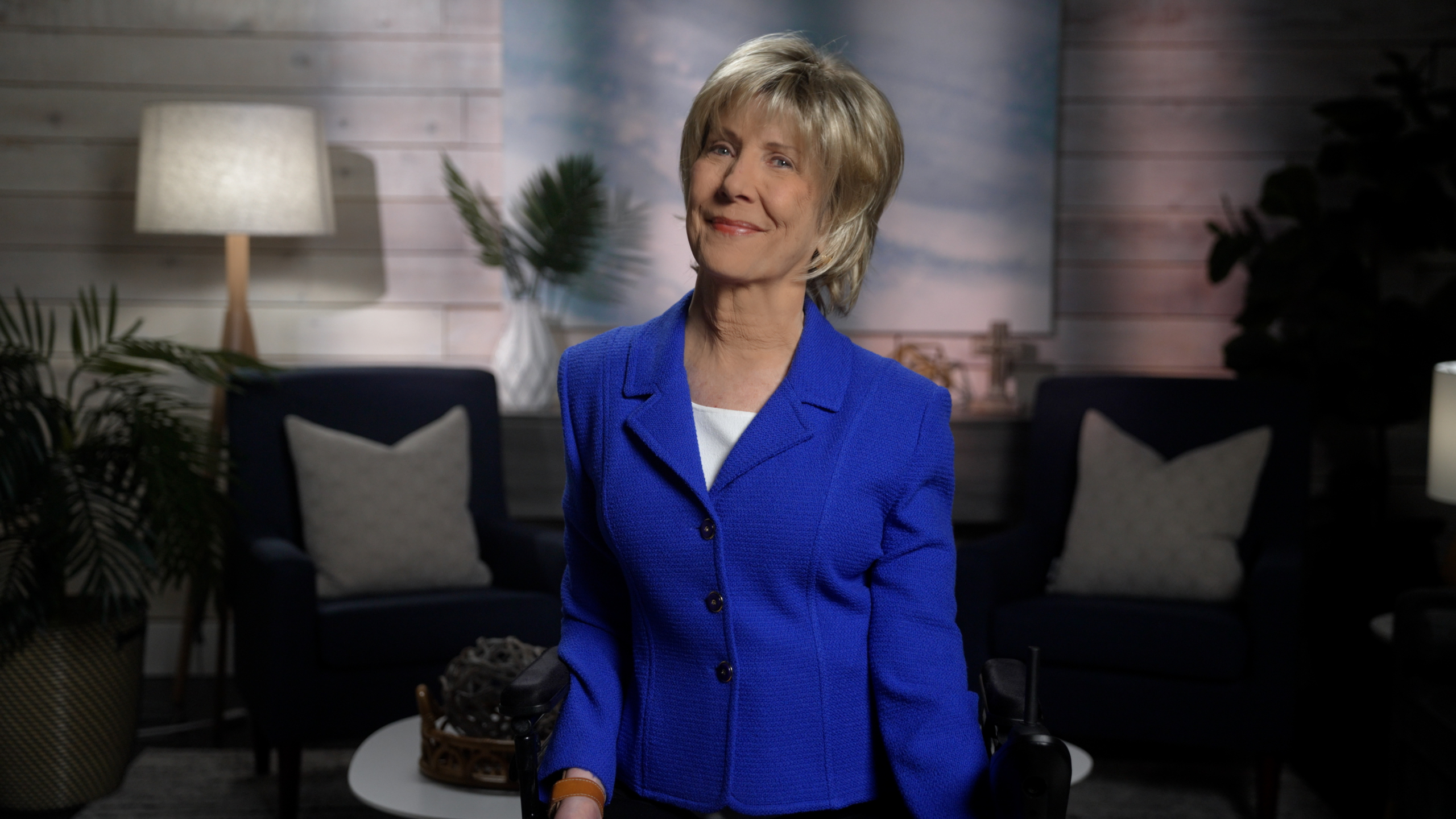 Joni Eareckson Tada is smiling for the camera, wearing a royal blue jacket over a white top. She is positioned in front of a softly blurred background that features a cozy living room setup.