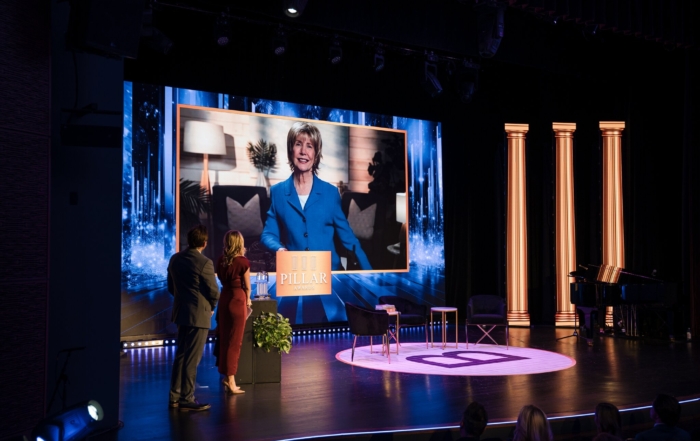 Joni Eareckson Tada on a large screen accepting an award in front of an audience in a large theater while a man and woman stand on the side of the stage facing the large screen where Joni is displayed via live video stream.