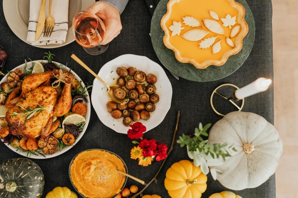 A table full of Thanksgiving dishes, a plate of whole turkey, a plate of mushrooms, mashed butternut squash, pumpkin pie, a glass of red wine, a lit up white candle, and pumpkins and flowers surrounding the table.